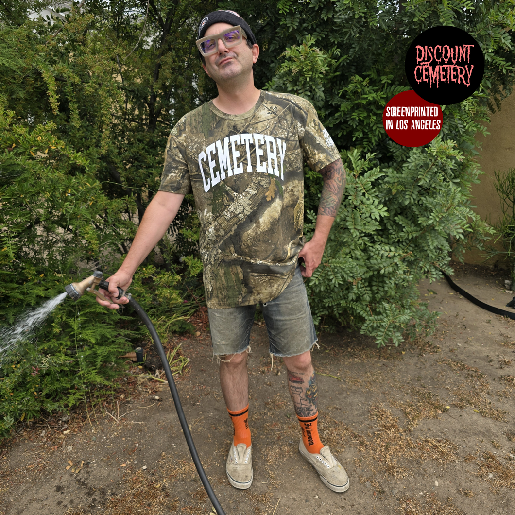 Man in real tree camouflage 'Cemetery' t-shirt holding a hose outdoors with 'Discount Cemetery' logo.