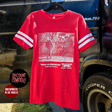 Load image into Gallery viewer, Red t-shirt with graphic design hanging on a hanger in front of a vehicle.